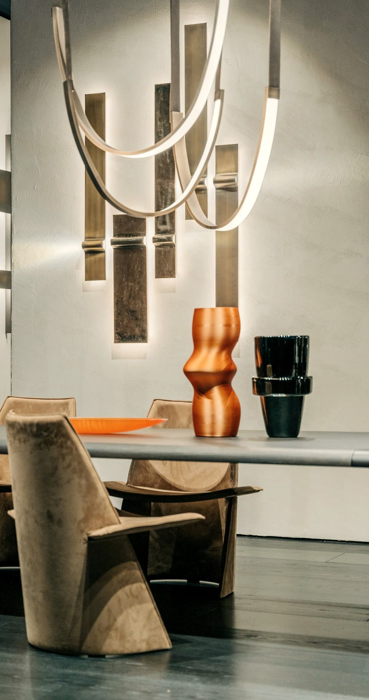 Backlit brass panels on a plaster wall above a dining table, with suede chairs and sculptural vessels